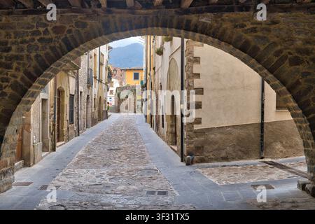 Vue à travers une arche de pierre ancienne encadrant une rue pavée étroite et sinueuse bordée de vieux bâtiments et de balcons à Anglès, Girona, Espagne Banque D'Images