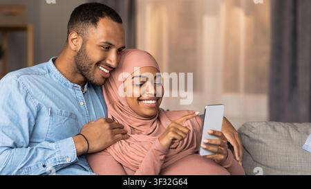 Femme afro-américaine enceinte heureuse et son partenaire souriant en utilisant un smartphone à la maison Banque D'Images