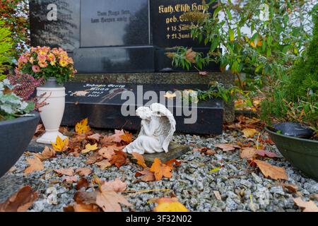Rossdorf, Allemagne - 20 octobre 2025 : Une petite figurine d'ange blanc repose parmi des feuilles d'automne colorées éparpillées sur du gravier devant des pierres tombales en marbre sombre Banque D'Images