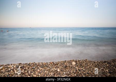 Photographie à longue exposition des douces vagues de la mer Méditerranée déferlant sur une plage de galets colorée. Banque D'Images