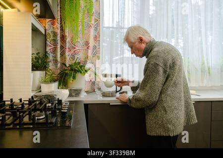 Vue de côté d'un senior voûté versant du thé dans une tasse. L'image évoque les défis et la dignité silencieuse de la vie quotidienne à la vieillesse. Grand-père se sert lui-même. Banque D'Images