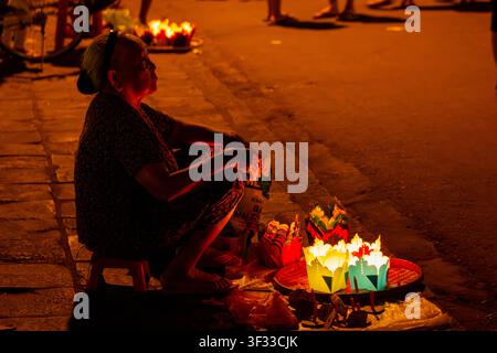 HOI AN, VIETNAM - 17 AOÛT 2015 : Une vendeuse de rue assise sur un tabouret bas vendant des lanternes illuminées à un étal de rue dans la vieille ville de Hoi An Banque D'Images