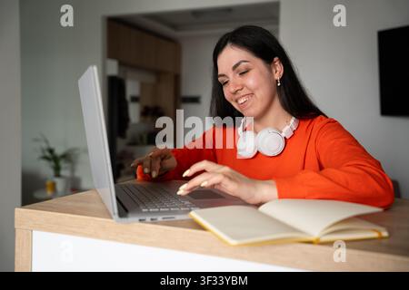 Jeune femme utilisant un ordinateur portable à son bureau à domicile avec un cahier ouvert et un casque d'écoute. Travail à distance, apprentissage en ligne et éducation à distance. Banque D'Images