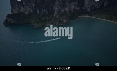Vue aérienne d'un bateau longtail laissant un sillage blanc sur la mer turquoise sombre près des hautes falaises de calcaire d'Ao Nang, Krabi, Thaïlande. Banque D'Images
