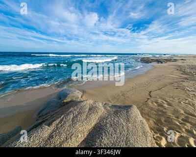 Sable doré rencontre des vagues turquoise sous un ciel bleu éclatant. Les vagues roulent doucement vers les rivages rocheux, laissant derrière elles des ondulations douces. Les nuages dérivent paresseusement Banque D'Images