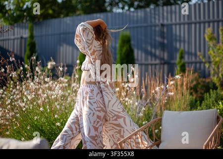 Jeune femme marchant dans un jardin d'été portant un ensemble de vêtements de maison élégants : robe, soutien-gorge, pantalon parmi les fleurs et les meubles de jardin. Banque D'Images
