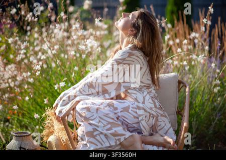 Jeune femme se détend sur une chaise dans un jardin d'été, portant une robe élégante et tenant un chapeau de paille. La dame respire, profite du soleil parmi les fleurs Banque D'Images