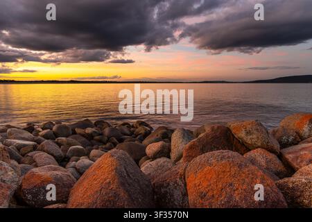 Vue du coucher de soleil sur l'océan avec de gros rochers couverts de lichen au premier plan et des nuages sombres dramatiques à Middleton Beach, en Australie-Occidentale, Austra Banque D'Images