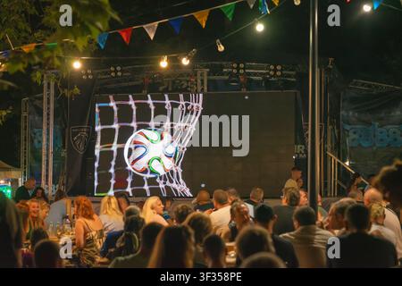 Bourgas, Bulgarie - 19 juillet 2026 : Des fans de football sont assis à des tables dans une zone de fans en plein air alors qu'une animation de football apparaît sur un grand écran LED avant le Banque D'Images