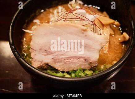 Close-up d'une soupe de nouilles Hokkaido authentique avec un bouillon riche, du ventre de porc tranché et une garniture d'oignon vert dans un bol sombre sur une table en bois. Banque D'Images