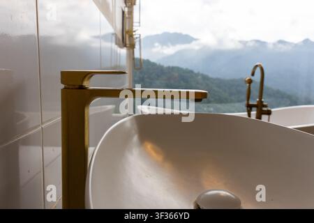 Un lavabo de salle de bain moderne avec robinet doré et vue sur les montagnes, un élégant bassin blanc et des accessoires métalliques chaleureux créant une atmosphère calme et luxueuse avec la nature Banque D'Images