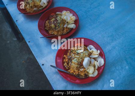 Assiettes de riz frit indonésien garni d'un œuf frit, de pâte de piment et de crackers krupuk, servis dans des assiettes en plastique rouge sur une table en métal. Banque D'Images