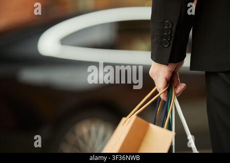 Gros plan d'un acheteur d'âge moyen portant des sacs de courses à côté d'une voiture, suggérant un achat haut de gamme et une mobilité des consommateurs urbains. Idéal pour le luxe retail, le commerce électronique, les visuels de campagne de vente Banque D'Images