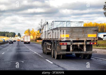 Véhicule industriel avec caisse ouverte circulant sur une route à plusieurs voies en journée, d'autres voitures et camions visibles au loin, arbres d'automne sur le bord de la route Banque D'Images