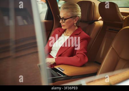 Femme d'affaires caucasienne d'âge moyen assise sur la banquette arrière d'une voiture, travaillant sur un ordinateur portable pendant le trajet, représentant la mobilité et la productivité des cadres. Vue latérale, idéale pour le marketing des voyages d'affaires Banque D'Images
