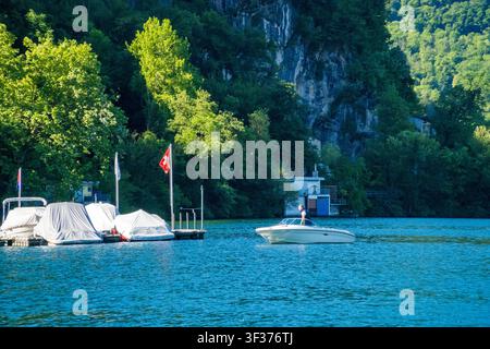 Caslano – Suisse – 12 mai 2026 : bateaux sur le lac de Côme avec drapeau suisse, falaise rocheuse et fond forestier, Malcantone, Tessin. Banque D'Images