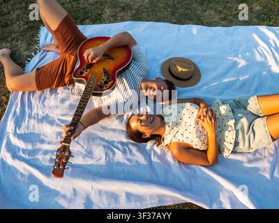 Un couple joyeux se détend sur une couverture de pique-nique dans un parc ensoleillé, riant ensemble tandis qu'une guitare acoustique repose à côté d'eux. Banque D'Images