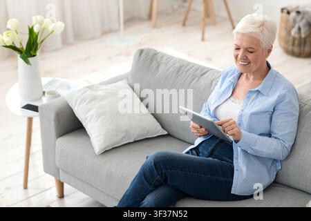 Femme moderne sénior se reposant sur un canapé à la maison avec un pavé numérique Banque D'Images