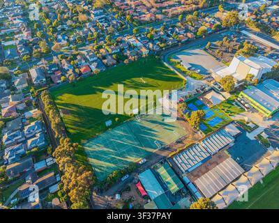 Une vue aérienne par drone capture une installation sportive suburbaine vibrante avec des terrains de jeu et des courts de tennis au milieu de maisons résidentielles calmes au coucher du soleil. Banque D'Images