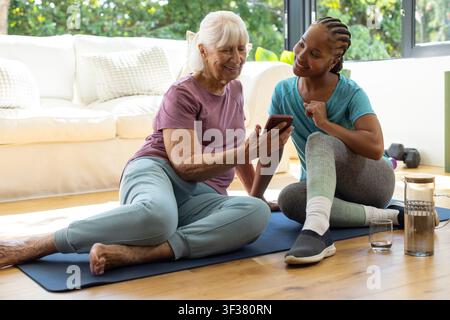 Coach féminine diversifiée en tenue de sport guidant une femme âgée sur des tapis de yoga à la maison en pointant un smartphone Banque D'Images