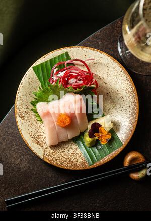 Vue par-dessus de tranches de sashimi de poisson blanc frais garnies de roe tobiko orange, magnifiquement disposées sur une assiette en céramique tachetée. Banque D'Images