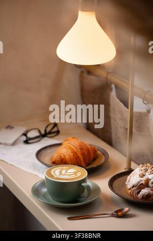 Plan en hauteur d'une table de café confortable éclairée par une lampe suspendue chaude, présentant une tasse verte de cappuccino avec un motif tulipe en latte art. Banque D'Images