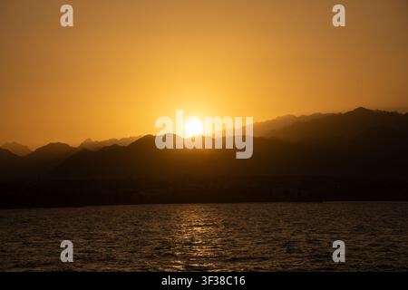 Soleil se couchant derrière une chaîne de montagnes, projetant une lumière chaude sur l'eau tranquille de l'océan Banque D'Images