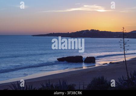 Le soleil se couche sur l'océan à Albufeira, projetant une lumière sur l'eau près d'une plage. Des rochers se trouvent sur le rivage tandis que les vagues clapotent doucement contre le sable. Banque D'Images