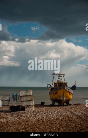 Seven Sisters, Hastings – 28.03.2026 – Le bateau de pêche jaune nommé Clara June est stationné sur une plage de galets près des casiers à homard sous un ciel nuageux au bord d'une mer calme. Banque D'Images