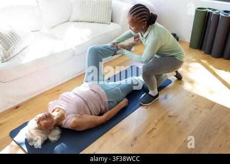Sénior en vêtements de sport recevant un étirement sur un tapis bleu à la maison, entraîneur afro-américain à genoux Banque D'Images