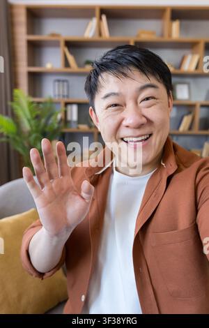 Homme asiatique heureux souriant et faisant un signe de la main lors d'un appel vidéo ou d'une réunion en ligne depuis son bureau à domicile, se connectant virtuellement avec des amis, la famille ou des collègues Banque D'Images