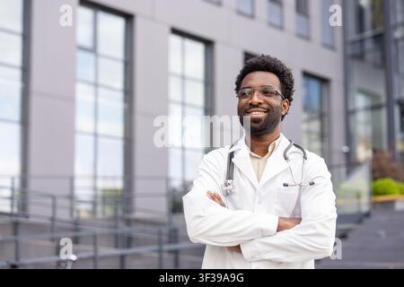 Médecin souriant debout les bras croisés, portant un stéthoscope et une blouse blanche, représentant le professionnalisme des soins de santé et l'expertise médicale en plein air Banque D'Images