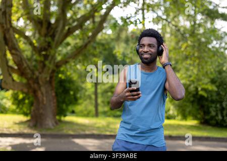 Homme souriant trouvant motivation et énergie dans la musique tout en s'adonnant à un entraînement physique en plein air, tenant un smartphone et portant des écouteurs sans fil pour un mode de vie actif dans un parc Banque D'Images