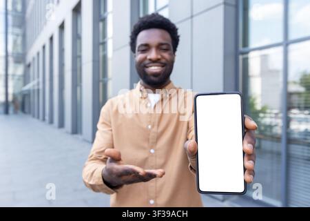 Jeune adulte souriant présentant un téléphone portable avec un écran blanc personnalisable vide, parfait pour mettre en valeur de nouvelles applications et du contenu numérique en plein air près d'un immeuble de bureaux Banque D'Images