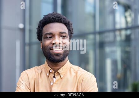 Jeune homme confiant avec une coupe afro souriant joyeusement tout en se tenant détendu à l'extérieur devant un bâtiment moderne, incarnant le succès, le bonheur et un avenir radieux Banque D'Images