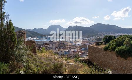 Vue panoramique de Carthagène, Espagne, avec des murs de pierre historiques, une skyline urbaine, des montagnes environnantes et un paysage méditerranéen sous un ciel bleu ensoleillé. Banque D'Images