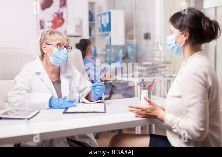 Docteur masqué consultant une jeune femme malade dans un bureau d'hôpital, montrant des soins cliniques avec des mesures de sécurité sanitaire protectrices, dans un cadre de consultation de santé. Banque D'Images