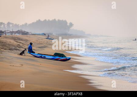 Bateau de pêche reposant sur la plage de sable au lever du soleil avec un pêcheur et un chien près de Chilaw sur la côte ouest du Sri Lanka Banque D'Images