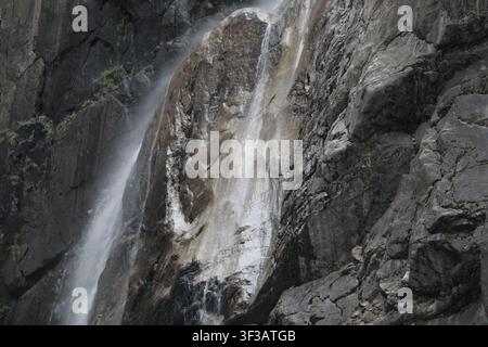 Une chute d'eau étroite cascade le long de falaises de granit abruptes, son ruban d'eau blanc contrastant avec la roche sombre et texturée sous un éclairage naturel dramatique. Banque D'Images