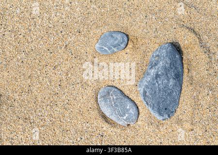 Trois pierres grises naturelles posées sur du sable doré de plage. Composition minimaliste mettant en avant des textures organiques et la lumière du soleil. Arrière-plan polyvalent Banque D'Images