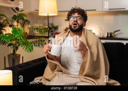 L'homme arabe à la maison se sent mal à la gorge tient tasse de thé chaud avec la médecine prend lente gorgée utilise le tissu. Un égyptien sur sofa cherche médecin sur smartphon Banque D'Images