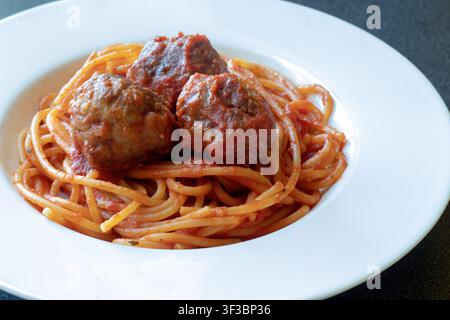 Gros plan incliné de spaghettis avec sauce tomate et trois boulettes de viande dans un bol blanc à large bord sur une surface sombre Banque D'Images