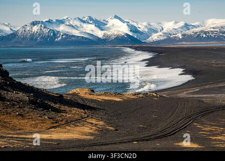 Une vaste plage de sable volcanique noir rencontre les vagues écumantes de l'océan en Islande. En arrière-plan, une majestueuse et longue chaîne de montagnes entièrement recouverte de neige Banque D'Images