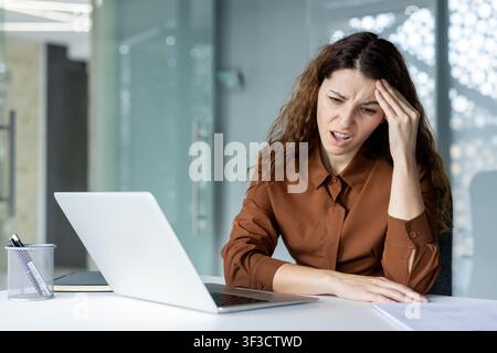 Jeune femme d'affaires subissant un violent mal de tête et un stress élevé en travaillant sur son ordinateur portable dans un bureau moderne, se sentant submergée par les responsabilités professionnelles et une charge de travail exigeante Banque D'Images