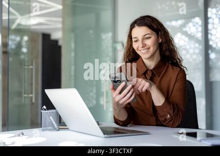 Jeune femme professionnelle interagissant joyeusement avec un smartphone moderne. Assise à un bureau blanc avec un ordinateur portable Banque D'Images