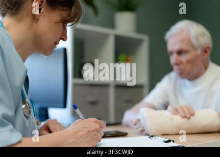 Médecin femme examinant un homme âgé écrivant sur un tableau dans une clinique. Le patient âgé reste calme tandis que la médecin prend des notes. L'homme âgé reçoit des soins alors que la médecin en blouse se concentre sur la documentation Banque D'Images