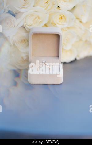 Bague de fiançailles solitaire en diamant dans une boîte en velours beige sur un fond de roses blanches douces, une nature morte romantique pour une demande en mariage Banque D'Images
