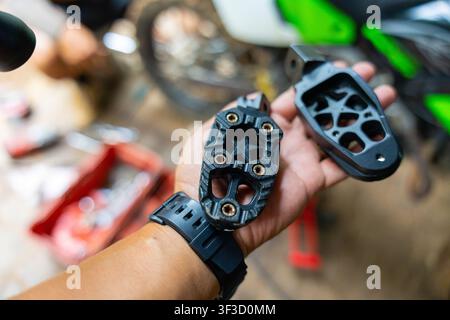 Une main tenant une paire de pièces de rechange pour repose-pieds de moto avec métal texturé et boulots de montage, fond d'atelier avec outils et moto créant f Banque D'Images