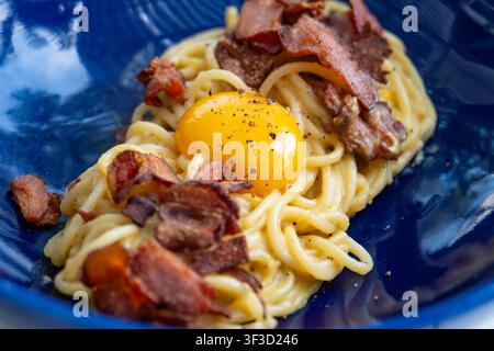 Close-up de spaghetti carbonara avec du bacon croustillant et un jaune d'œuf cru sur le dessus, servi dans une assiette bleue texturée Banque D'Images