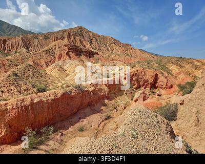 Collines érodées colorées et formations de grès rouge dans le canyon du Conte, au Kirghizistan. Couches géologiques, terrain sec et crêtes montagneuses sous un ciel bleu Banque D'Images